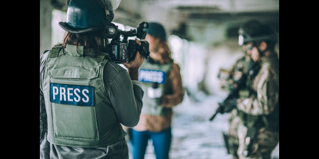 Female war correspondent wearing a hard hat pointing a camera at another correspondent in a war zone